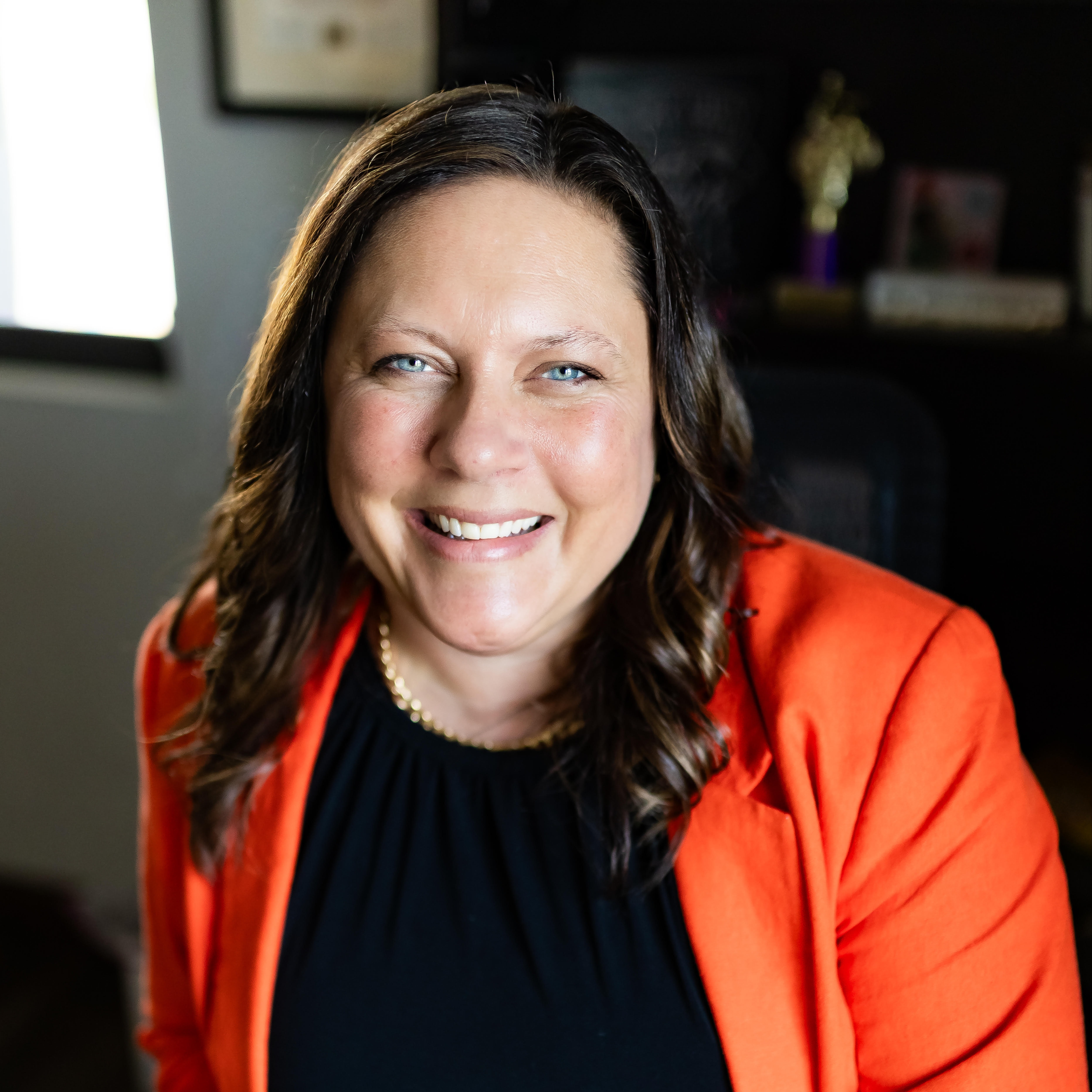 Woman with brown hair wearing a black top and orange blazer, smiling. A window and frame are visible in the background.