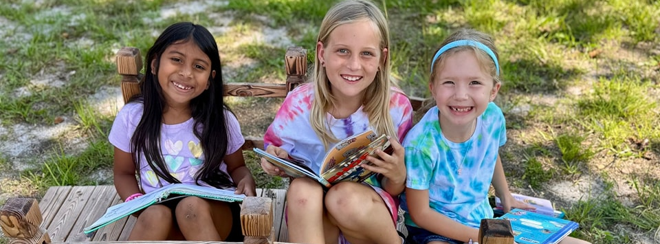 three young girls smiling outside and reading books