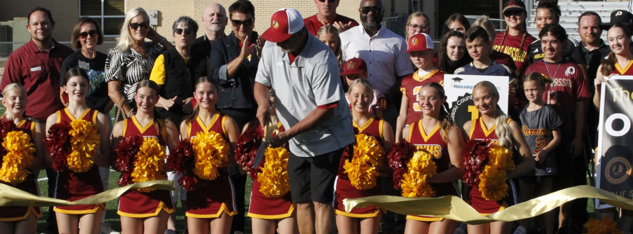 West Nassau High School ribbon-cutting ceremony held on the athletic field. Cheerleaders in maroon and gold uniforms kneel in the front holding pom-poms as a man cuts a large gold ribbon. Students, school staff, district representatives, and community members stand behind them to celebrate the occasion.