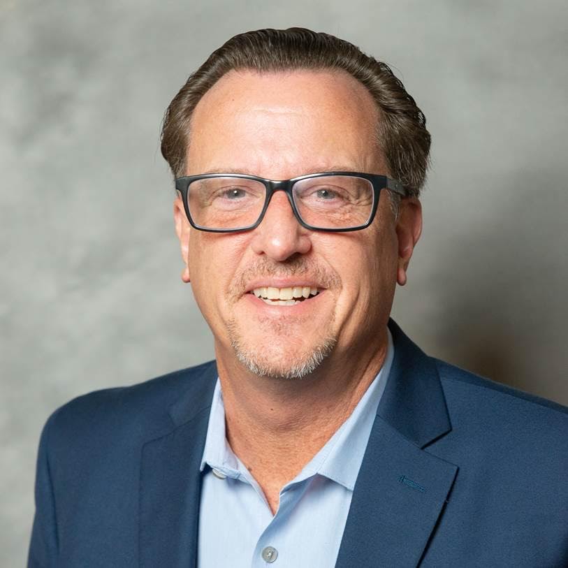 1. A man wearing glasses, a blue shirt, and a blue jacket stands confidently, smiling at the camera. 2. A man with glasses dressed in a blue shirt and jacket poses for the photo, exuding a professional appearance. 3. A man in a blue shirt and jacket, wearing glasses, looks directly at the camera with a friendly expression.