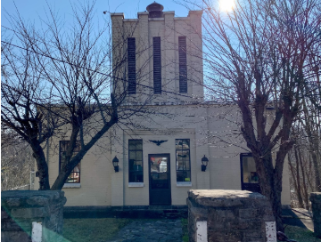 A historic cream-colored brick building with a prominent central tower featuring tall vertical windows. The building is framed by leafless trees and stone gateposts, with lantern-style lights flanking the double front doors. The sun shines brightly in the upper right corner of the image.