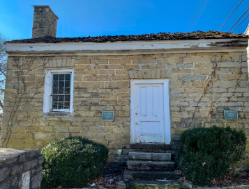 Front view of a small historic limestone building with a weathered white wooden door, a single-paned window with bars, and two informational plaques on the wall. The structure is framed by neatly trimmed bushes, stone steps, and a clear blue sky overhead.