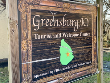 Decorative wooden sign for the Greensburg, Kentucky Tourist and Welcome Center, featuring a vintage-style frame, a green silhouette of Kentucky with an orange star marking Greensburg, and text noting sponsorship by FBLA and the Youth Action Council.
