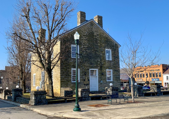 A tan stone building, the Old Greensburg Courthouse, features white-trimmed windows and a central white door. A green streetlamp stands in front, with bare trees and a blue sky in the background, hinting at early spring.