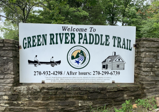 Sign marking the entrance to the Green River Paddle Trail, showcasing activities like canoeing and wildlife watching, with contact information below. The sign is set against a backdrop of trees and a stone wall.