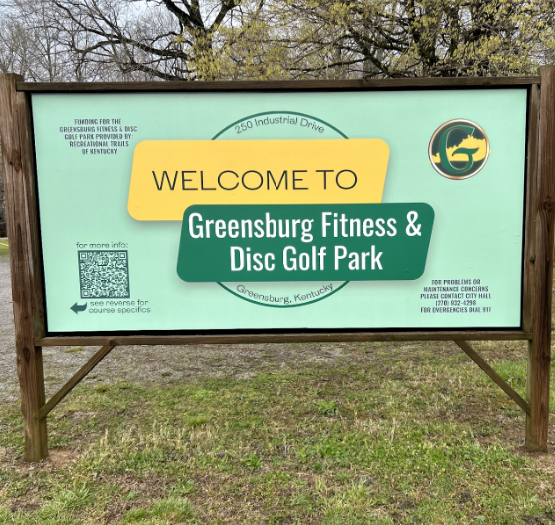 The Greensburg Fitness & Disc Golf Park welcome sign, a light green board with bold text, invites visitors to the free 9-hole course in Greensburg, Kentucky, and includes contact details and a QR code.