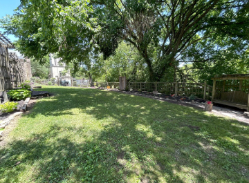 Grassy area in the Heritage Garden with trees, a wooden fence, and visible buildings beyond.