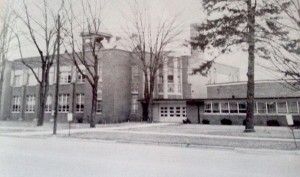 A large, multi-story building with a garage and trees in front, seen from across the street on a cloudy day.