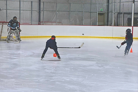 two kids on the ice shooting a puck at a hockey goalie in front of them with hockey sticks. 
