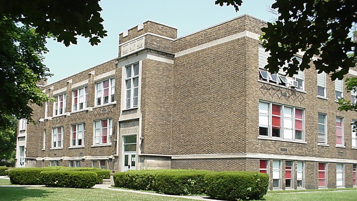 A brick school building with multiple windows and a green entrance, surrounded by neatly trimmed bushes and trees.