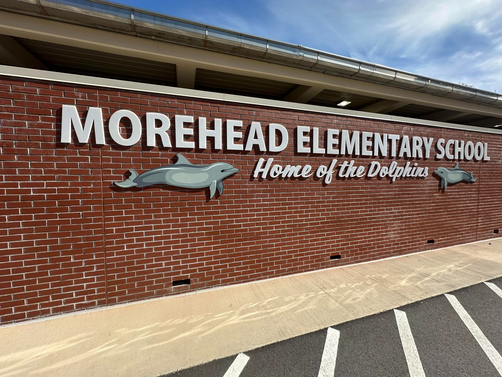 Image of a brick wall with a sign that reads Morehead Elementary School Home of the Dolphins with 2 dolphin icons on either side of the sign.