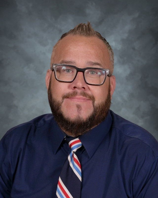 A man with glasses and a beard, dressed in a blue shirt and tie, poses for the camera.