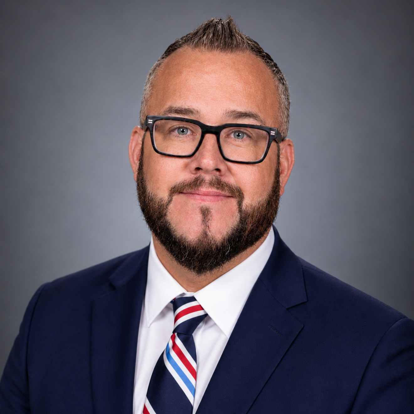 A man with glasses and a beard, dressed in a blue shirt and tie, poses for the camera.