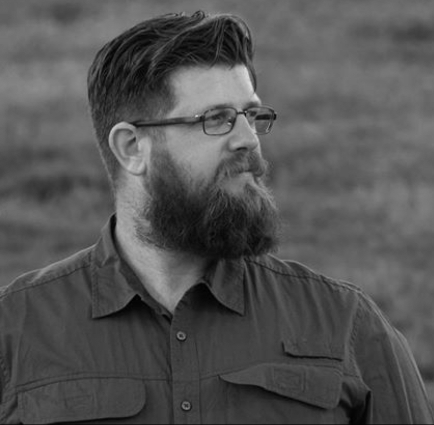 A bearded man wearing glasses stands in front of a lush green field under a clear blue sky.