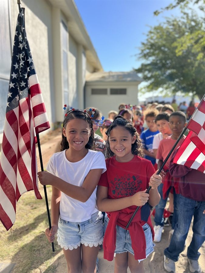 Two young girls proudly hold American flags in front of a building, celebrating patriotism and unity.