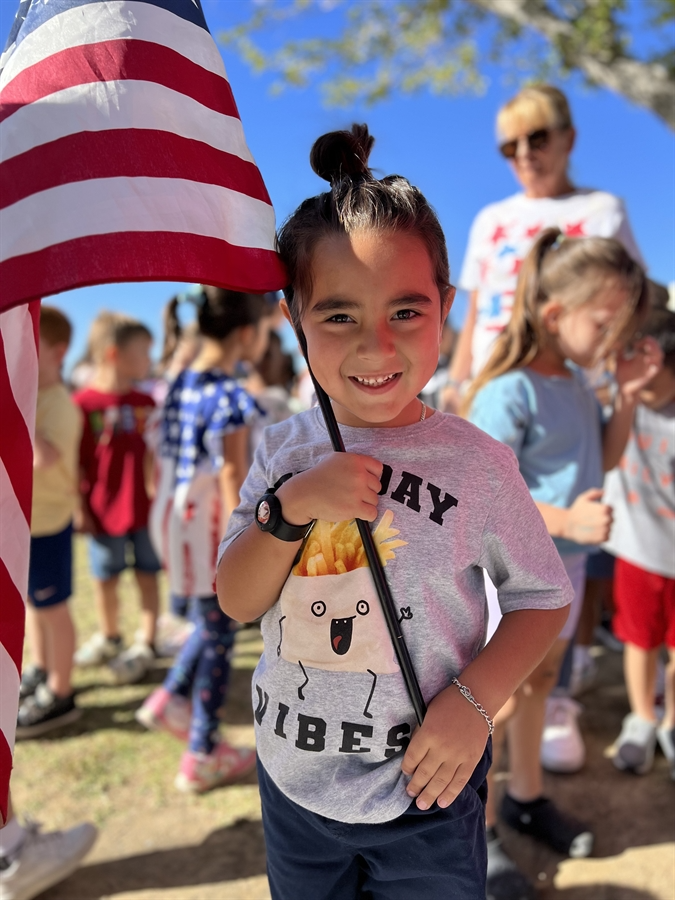A young girl proudly holds an American flag, smiling and standing against a bright blue sky.