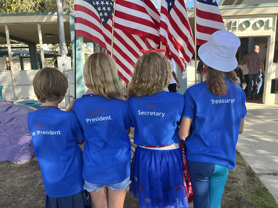 A young girl proudly holds an American flag, smiling and standing against a bright blue sky.