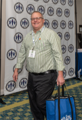 1. A man wearing a green shirt and blue pants is holding a blue bag.   2. A man in a green shirt and blue trousers holds a blue bag.   3. A man dressed in a green shirt and blue pants carries a blue bag.  
