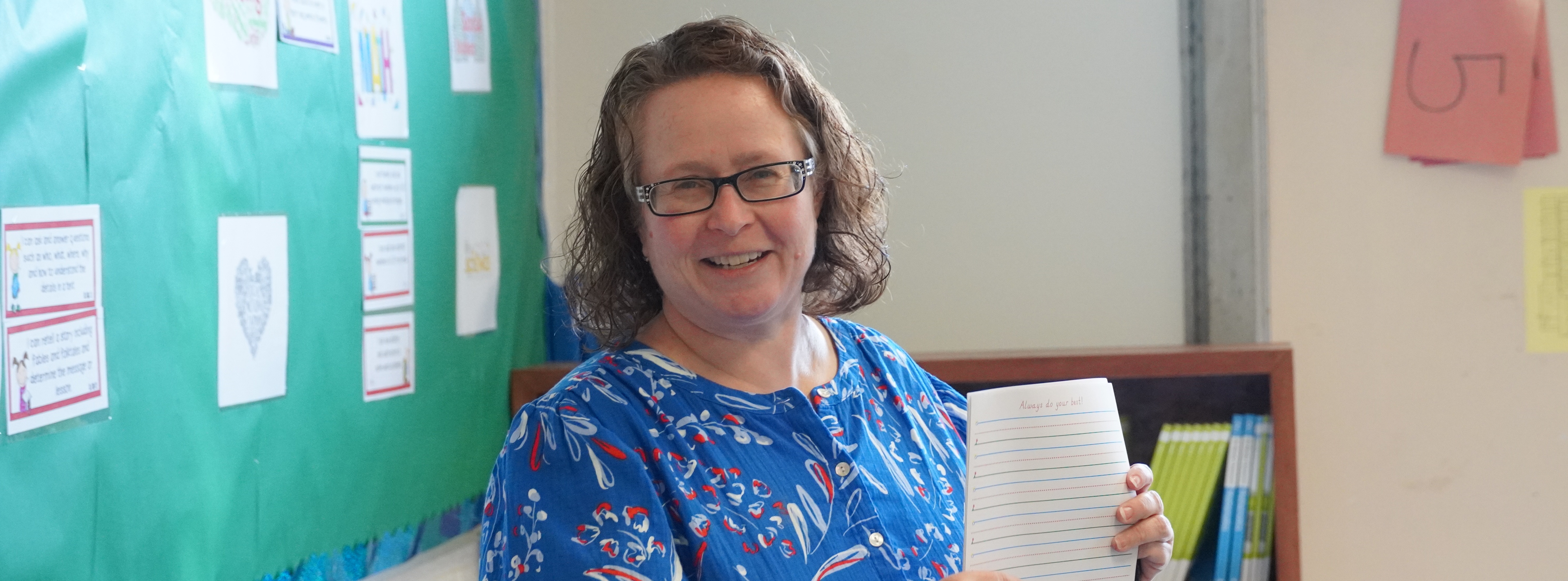 Teacher stands in a classroom holding up a lined writing sheet, smiling toward the camera, with student materials and a bulletin board visible behind her.