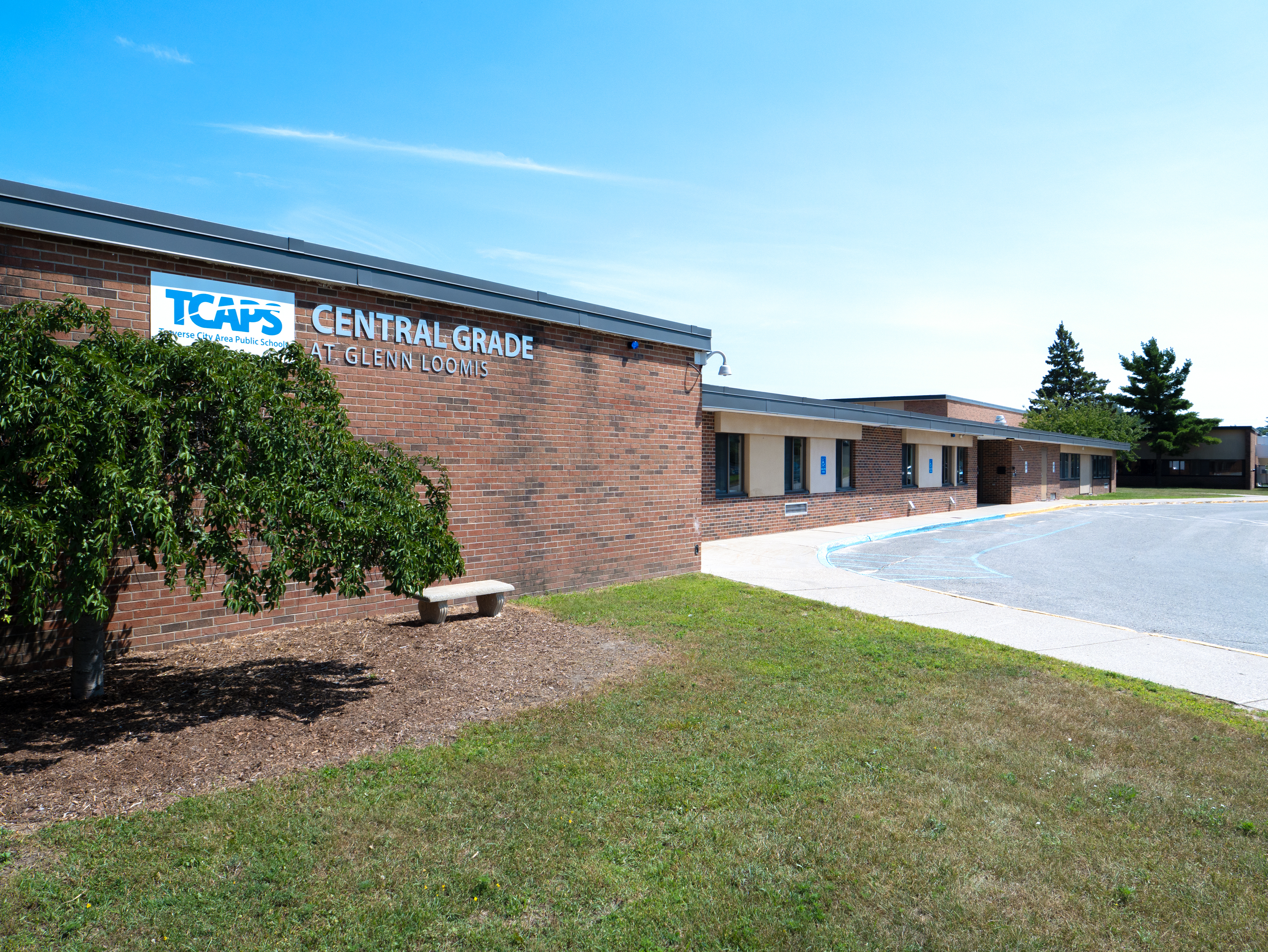 A building with "TCAPS CENTRAL GRADE SCHOOL" sign, a bench, and a grassy area. The sky is blue.