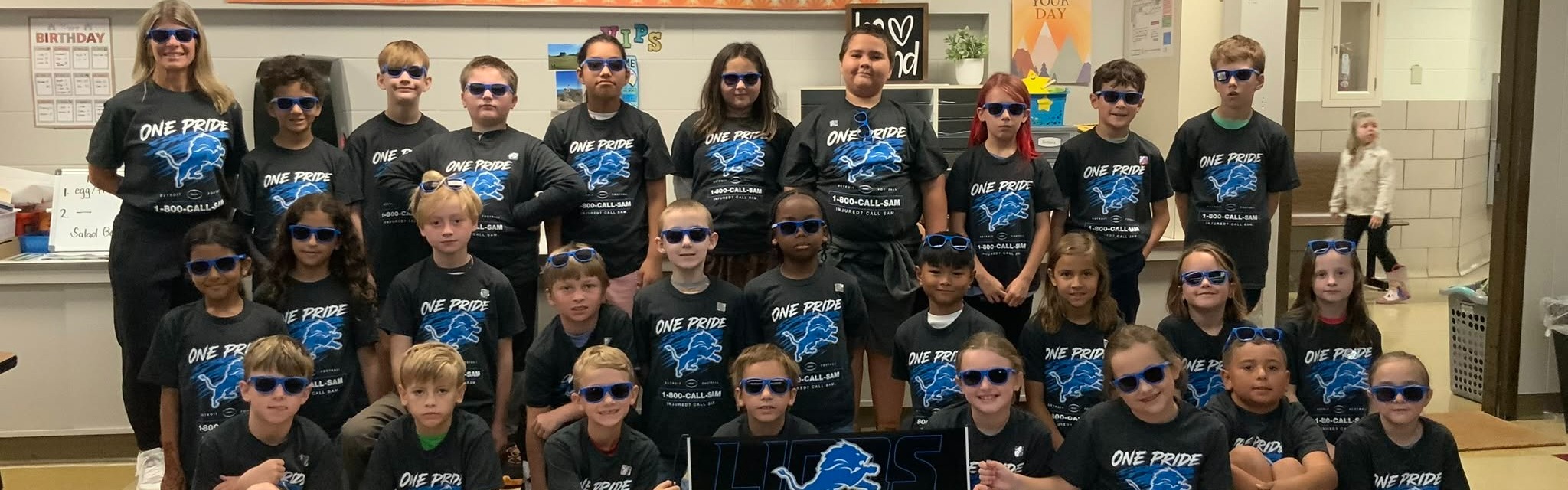 Group of young individuals in matching blue shirts and sunglasses, posing for a photograph in a classroom.
