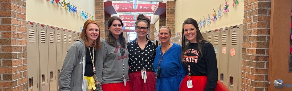Five women in tutus and boots stand in a hallway. They have their arms crossed, and a locker line is behind them.