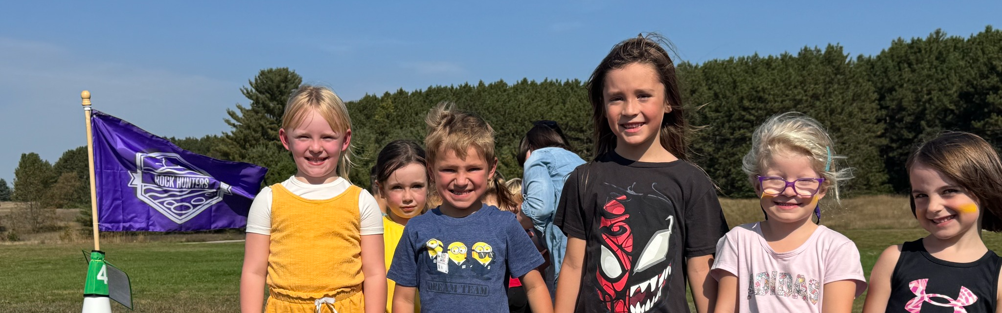 Six young children smiling and holding hands in a field with a purple school flag. 