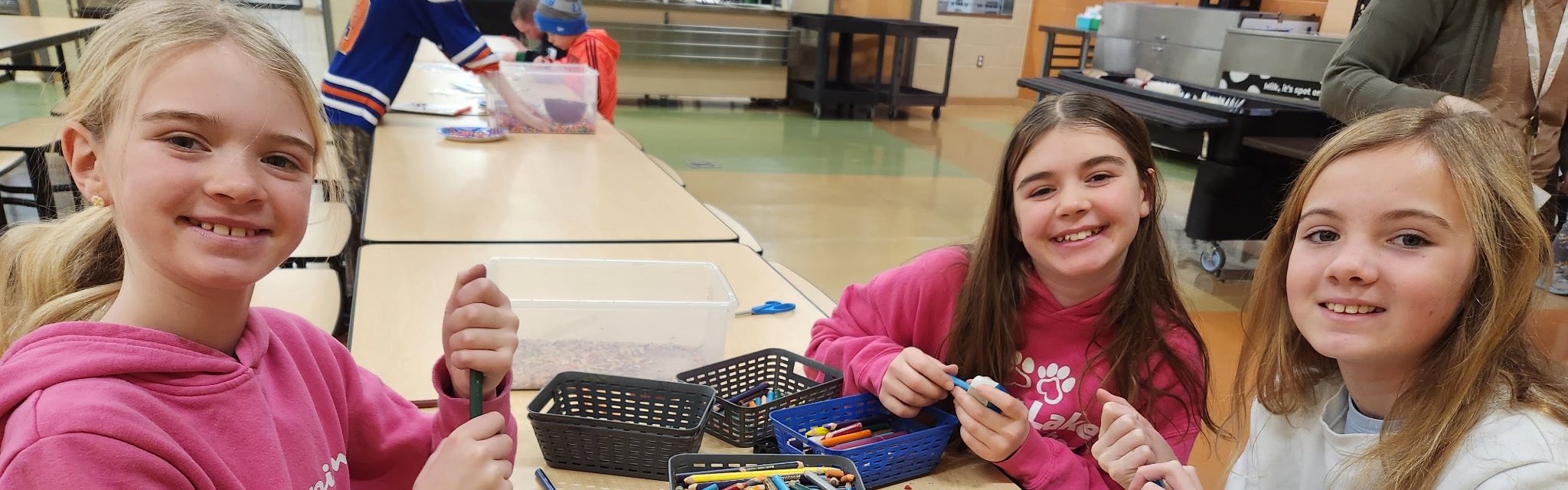 Three girls sit at a table with colored pencils and baskets, smiling. Behind them, a room with tables and chairs.