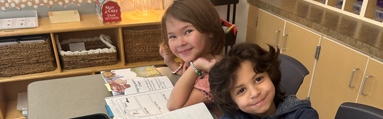 Two children sit at a desk with papers and a book. One holds a pencil, and the other smiles.