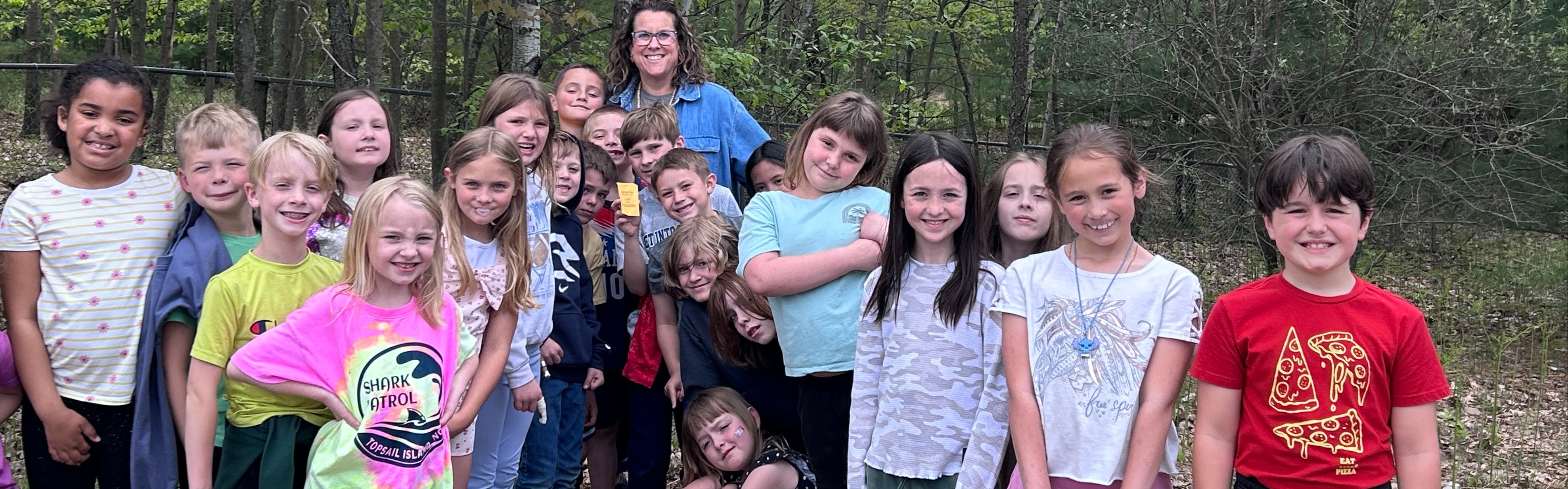 A group of children in a wooded area, wearing various clothing. They are gathered around a woman.