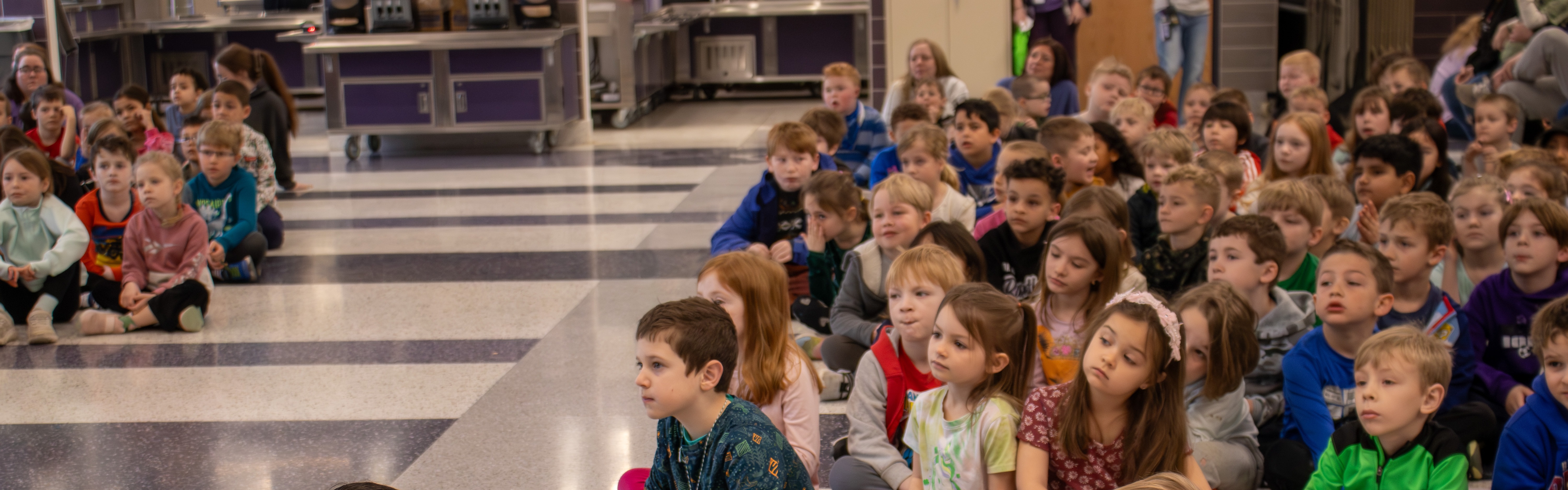 A group of young students sit on the floor and listen intently during an assembly.