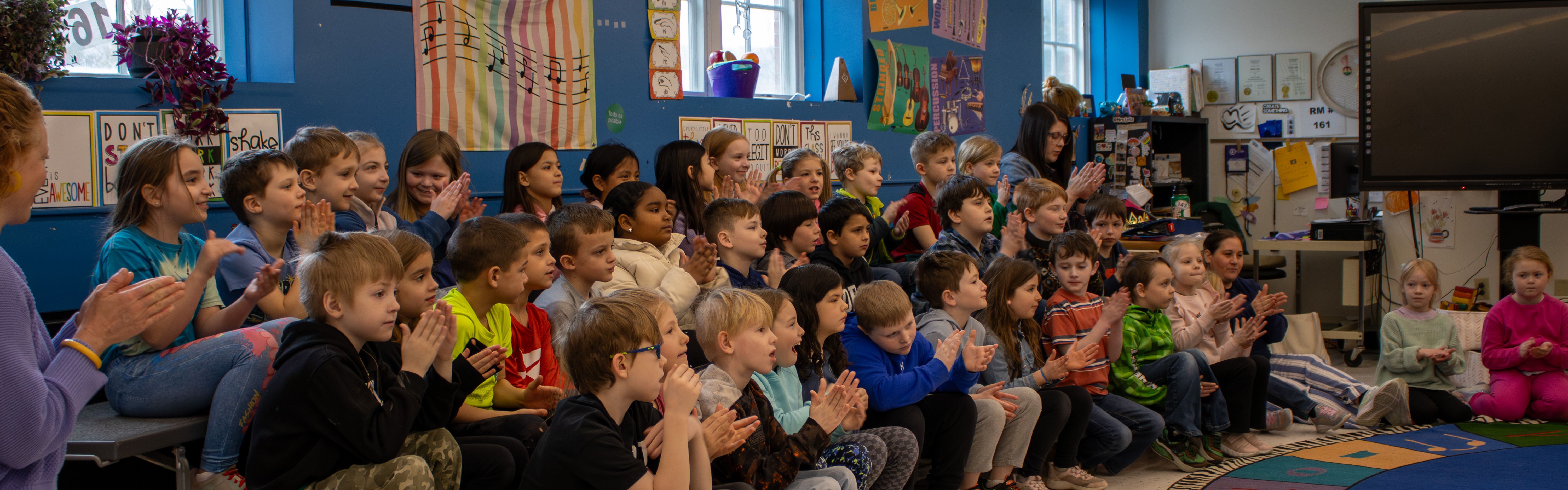 Students clap excitedly while sitting on bleachers in a music classroom. 