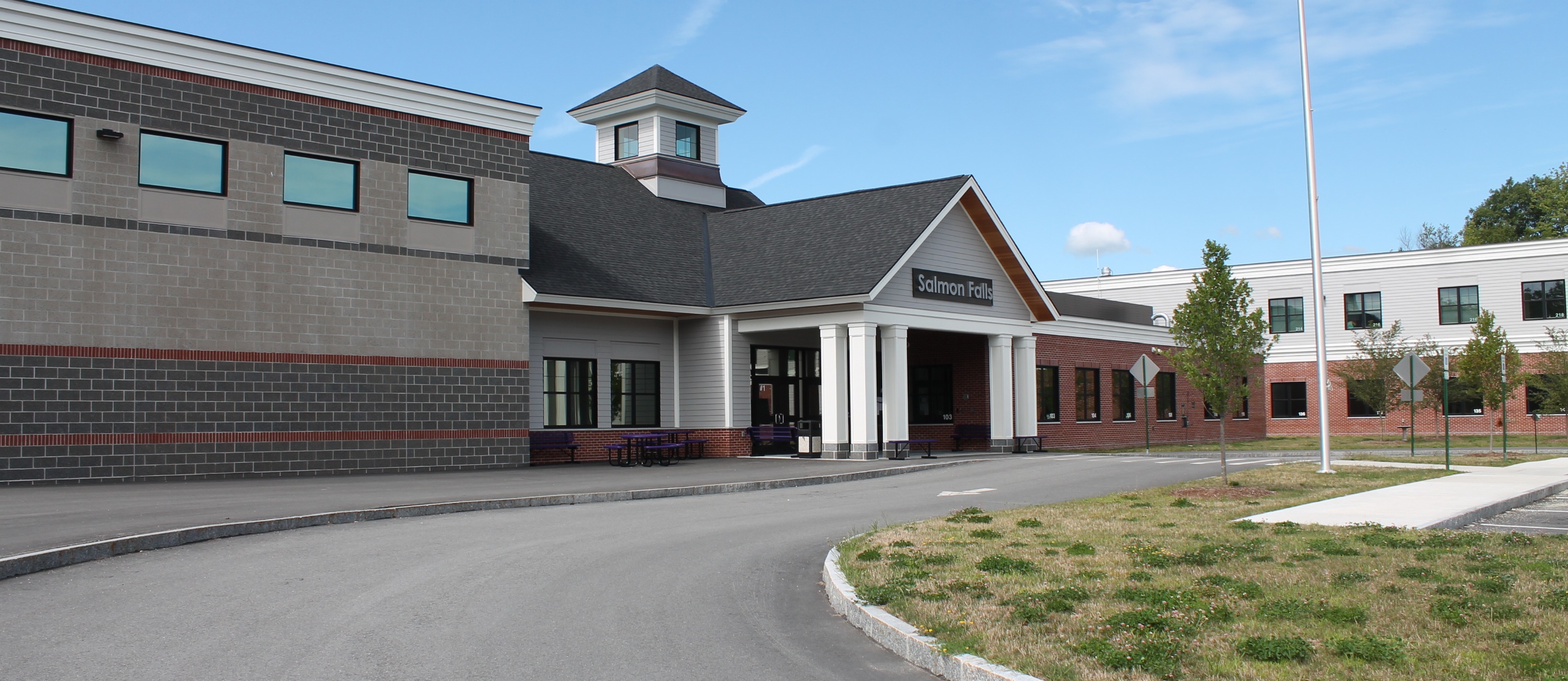Road with Salmon Falls School on the left, featuring windows and a tower, and a grassy area. Clear sky.