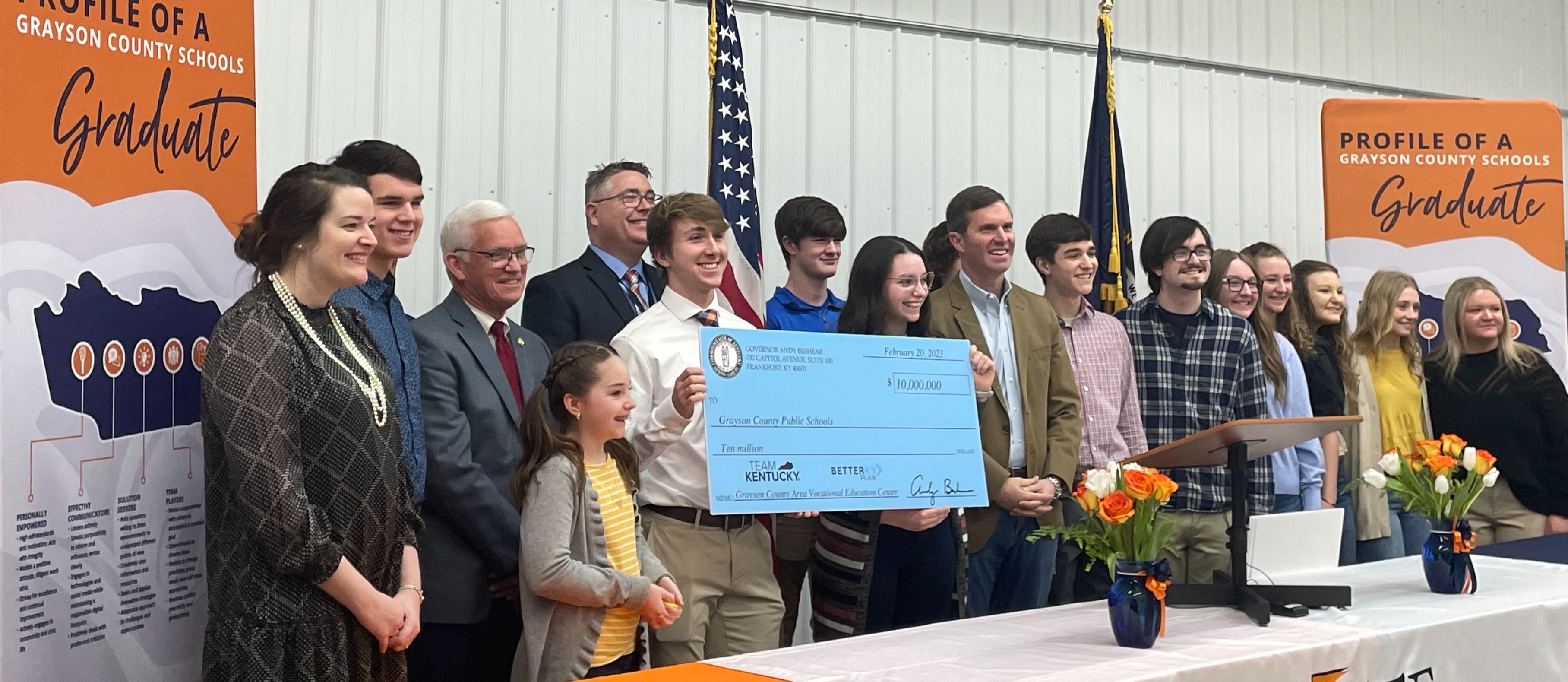 GCS students, Supt Doug Robinson with Gov. Beshear , Steve Meredith and Samara Heavrin holding a large ceremonial check standing in front of an orange, white and navy draped table with CTE logo. American and KY flags behind and flanked by Profile of a GCS graduate vertical banners.