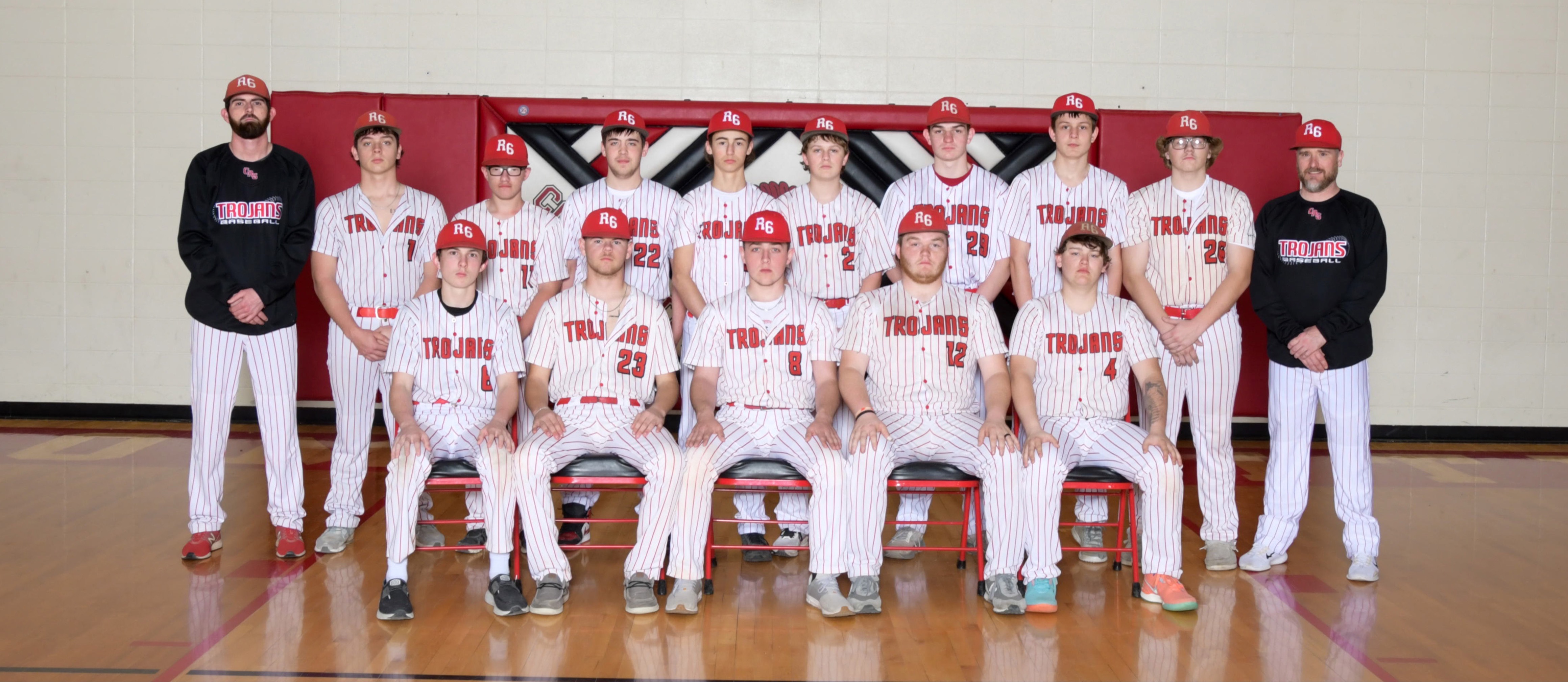 A high school baseball team in red and white uniforms poses for a group photo in a gymnasium under a basketball hoop.