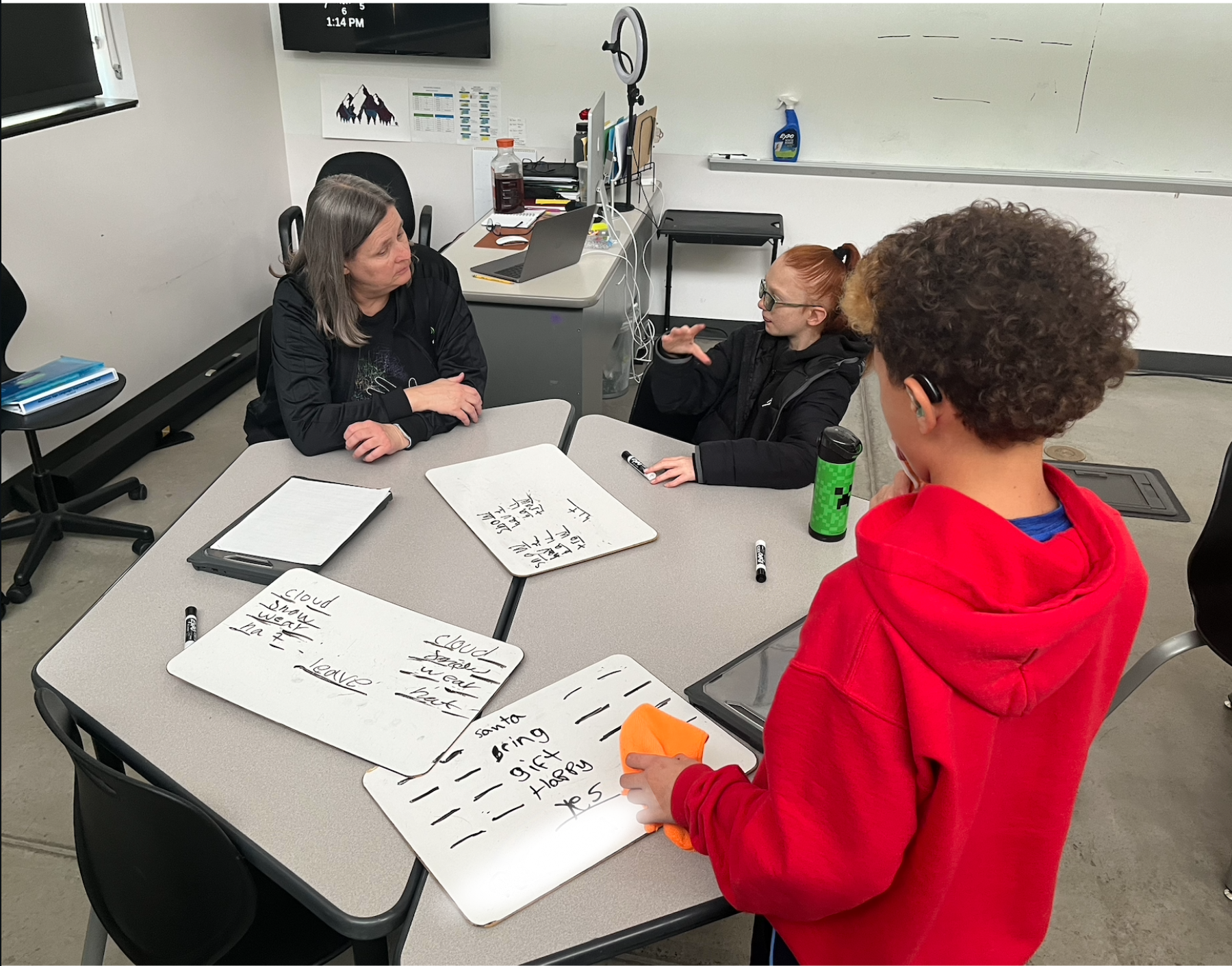 Two students are looking at their teacher, there are white boards on the table with words written for a class activity.