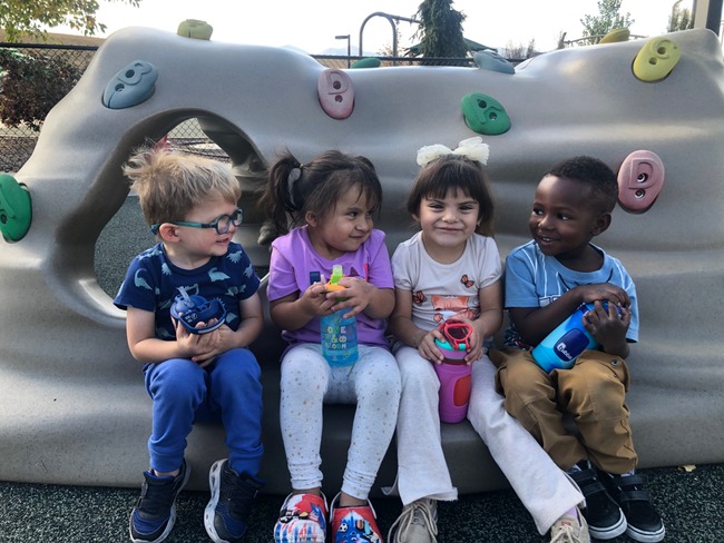 Four preschool students are sitting on a little rock climbing structure, smiling and laughing at each other.