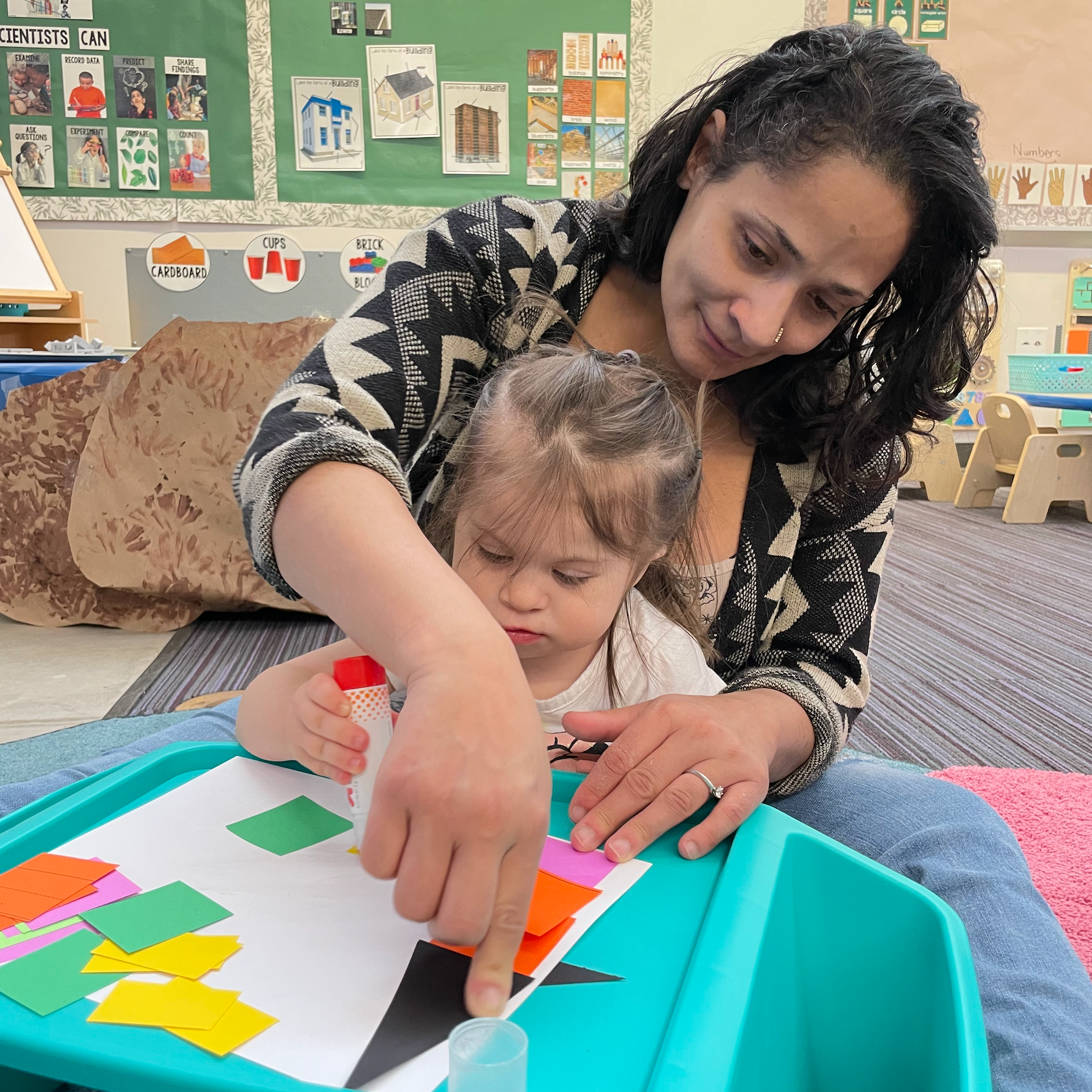 This image shows an adult and a child engaged in a hands-on activity, likely in a classroom or daycare setting. The adult is helping the child glue colorful pieces of paper onto a white sheet, possibly as part of an arts and crafts project. The background displays educational posters about science, building structures, and other learning materials, suggesting this is an early childhood education environment.