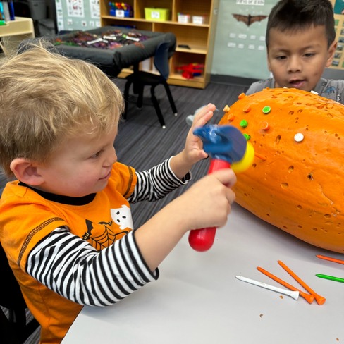 This photo shows two young children engaging in a hands-on activity—using toy hammers and colorful plastic pegs to decorate a large pumpkin. It captures a moment of focused fine motor development and creative play, likely part of a sensory or seasonal learning activity.