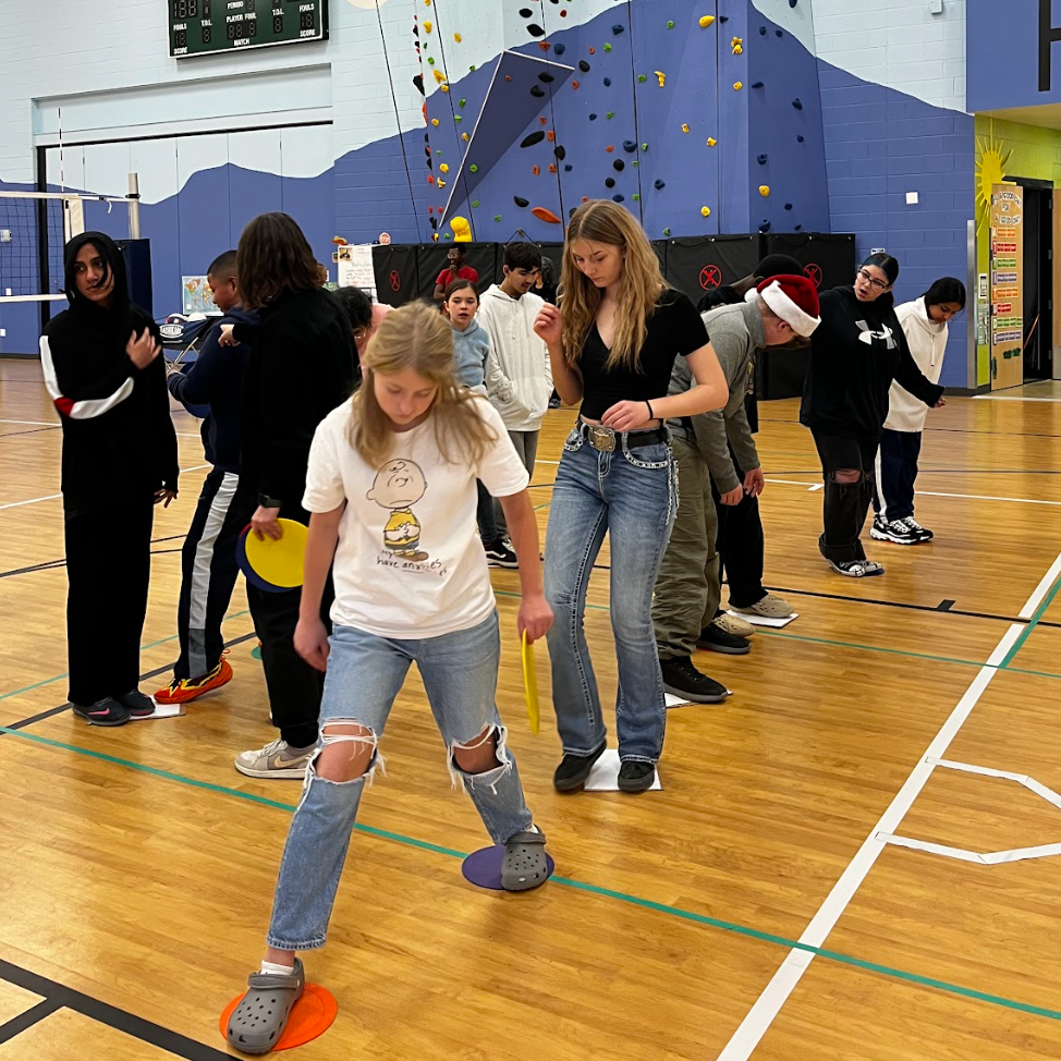 A group of middle and high school students participate in a team-building activity in a gymnasium, stepping on colored discs in a line. The background features a rock climbing wall and volleyball net, with a scoreboard and painted mountain mural on the wall.
