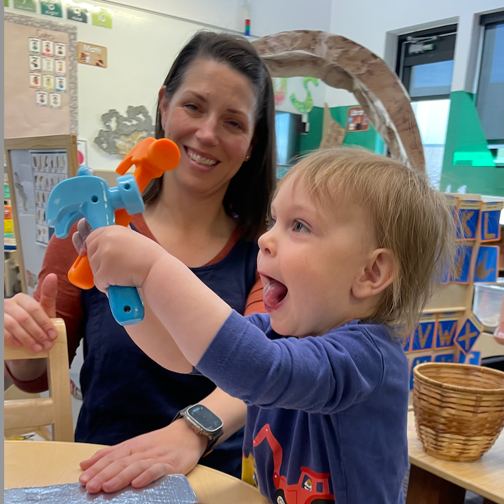 A smiling toddler enthusiastically holds up a toy hammer while sitting at a classroom table, with his parent nearby watching supportively.
