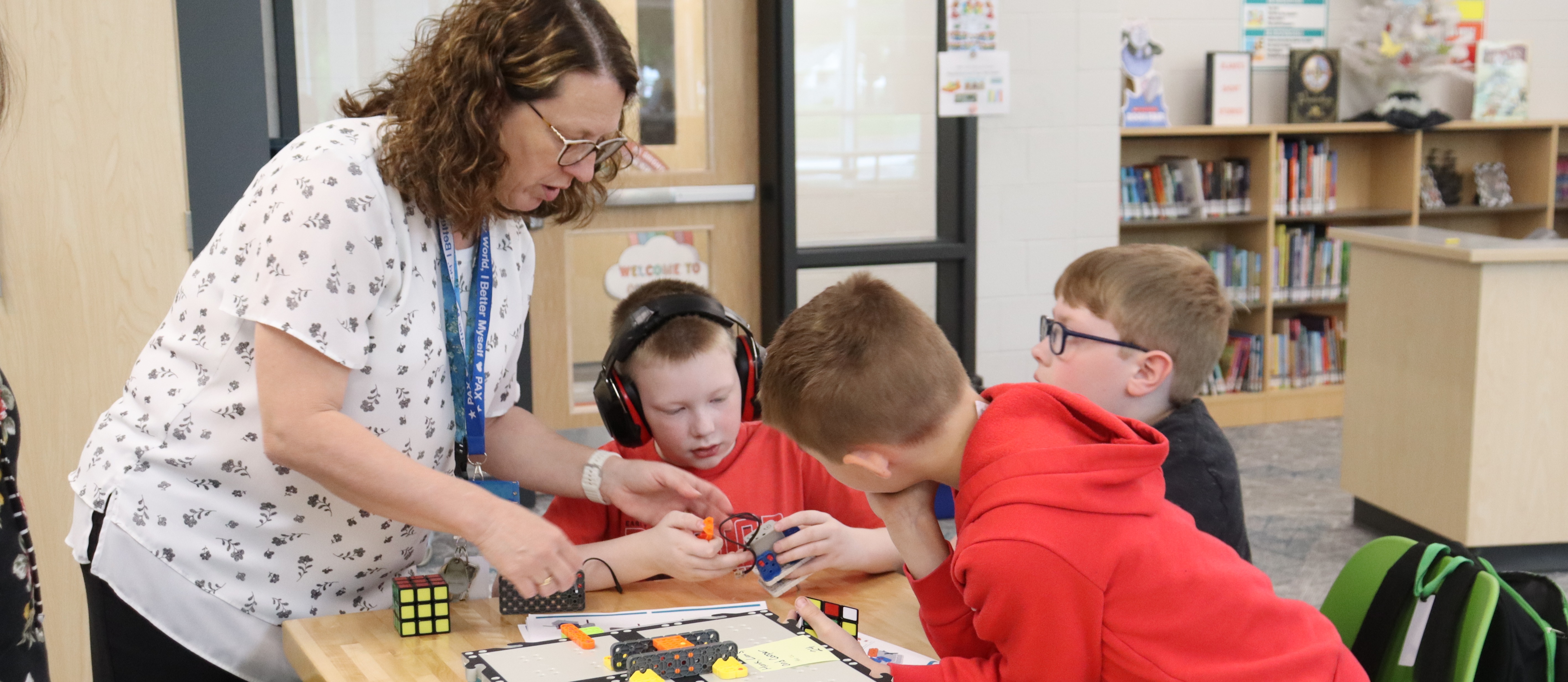 A woman and three children are at a table with a toy, one wearing headphones. A bookshelf is in the background.
