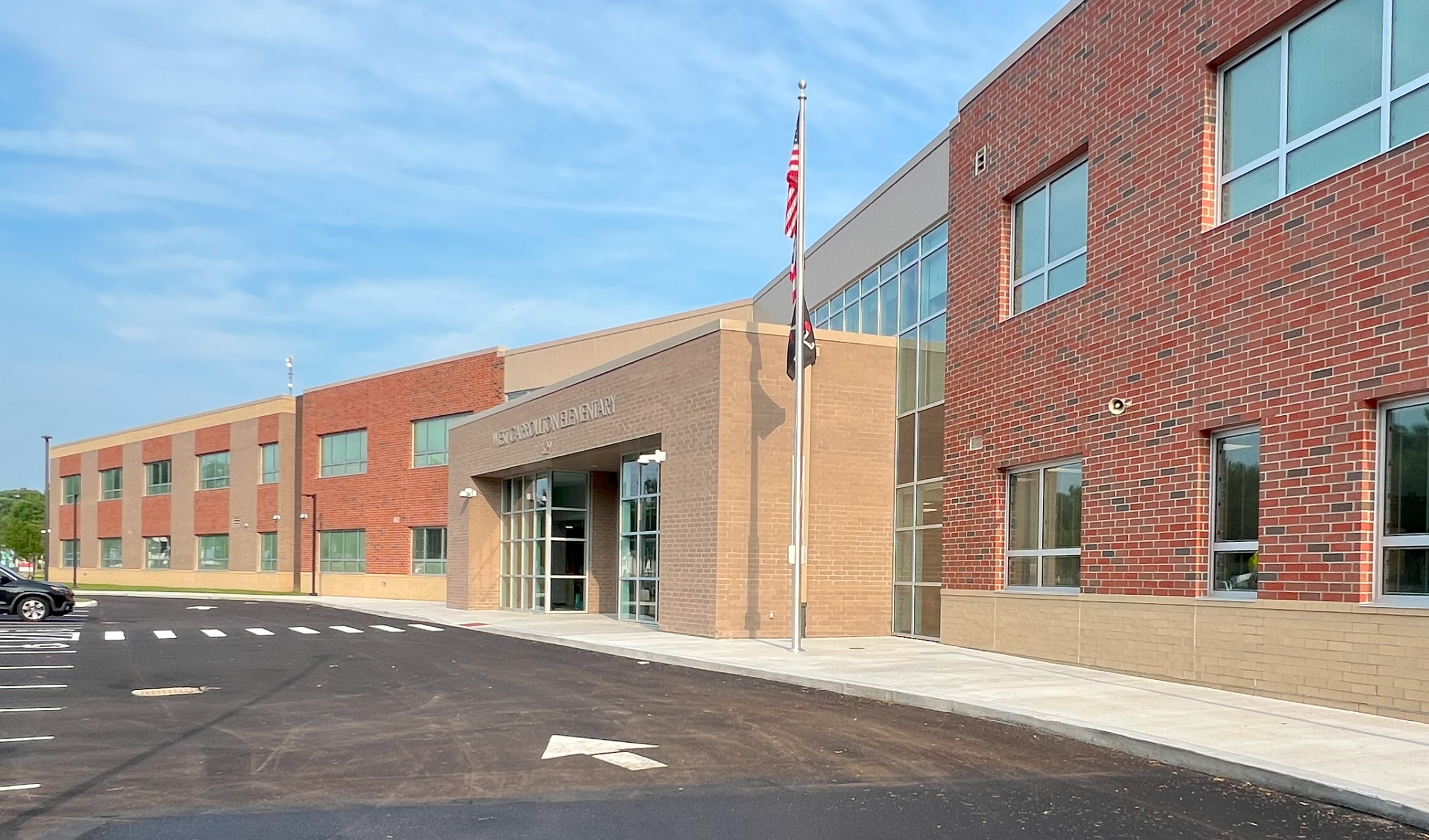 A school with brick walls, glass windows, and a flagpole in front of it. An empty parking lot is in front of it.