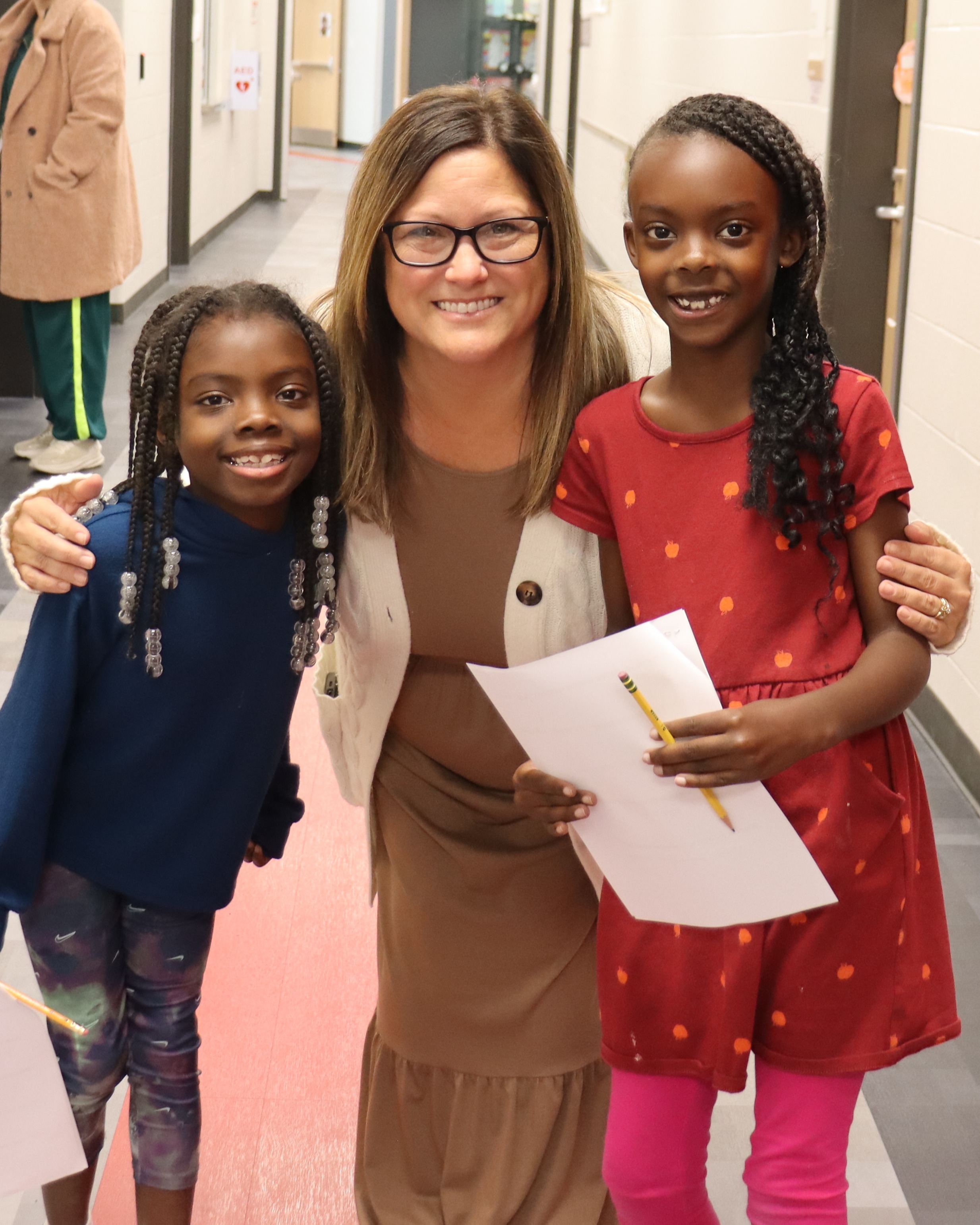 Woman and two girls in hallway. One girl holds paper and pen, other holds paper. Background includes doors and hallway lights.