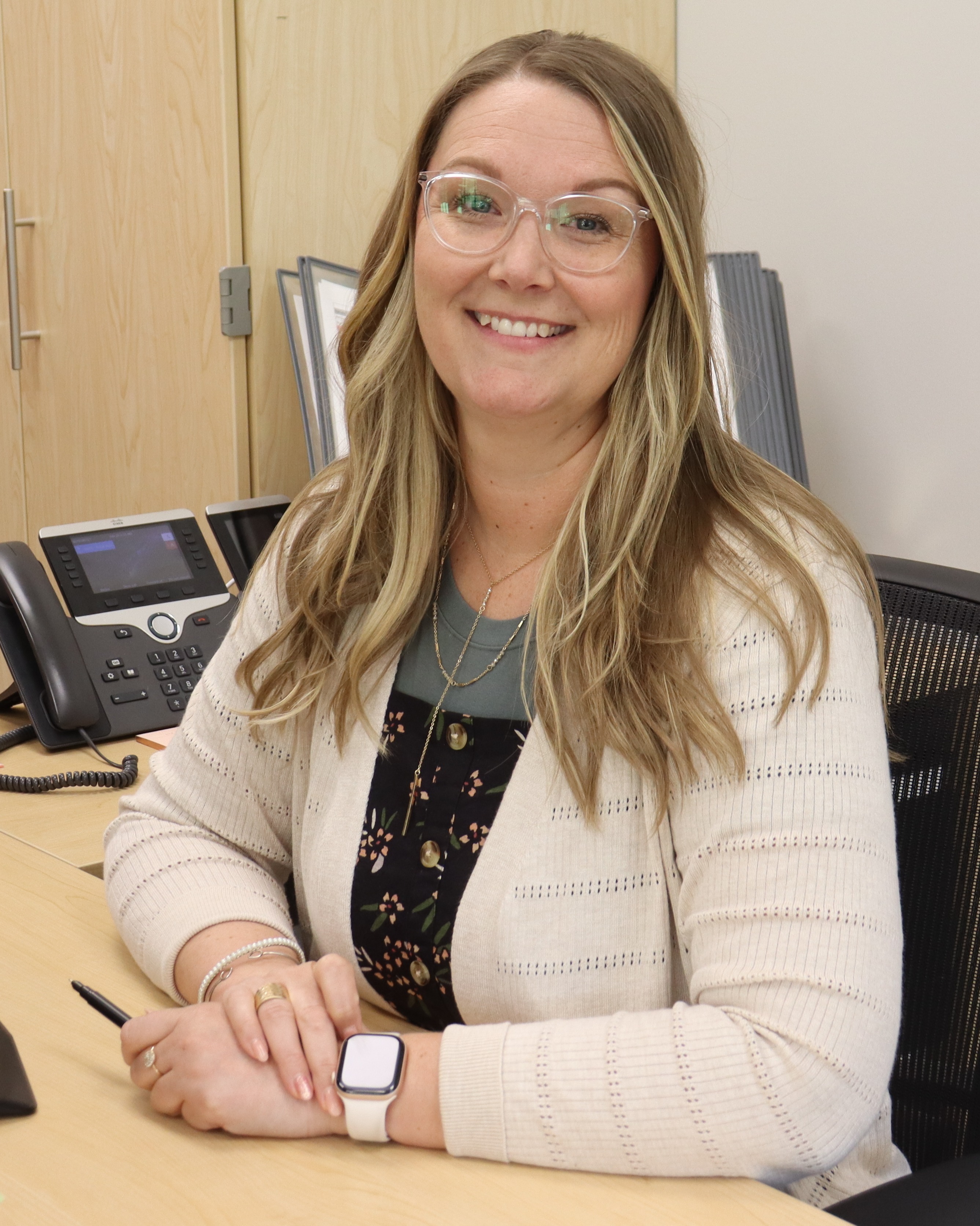 A woman sits at a desk with a computer and other items, smiling. Behind her is a wall with a power outlet.