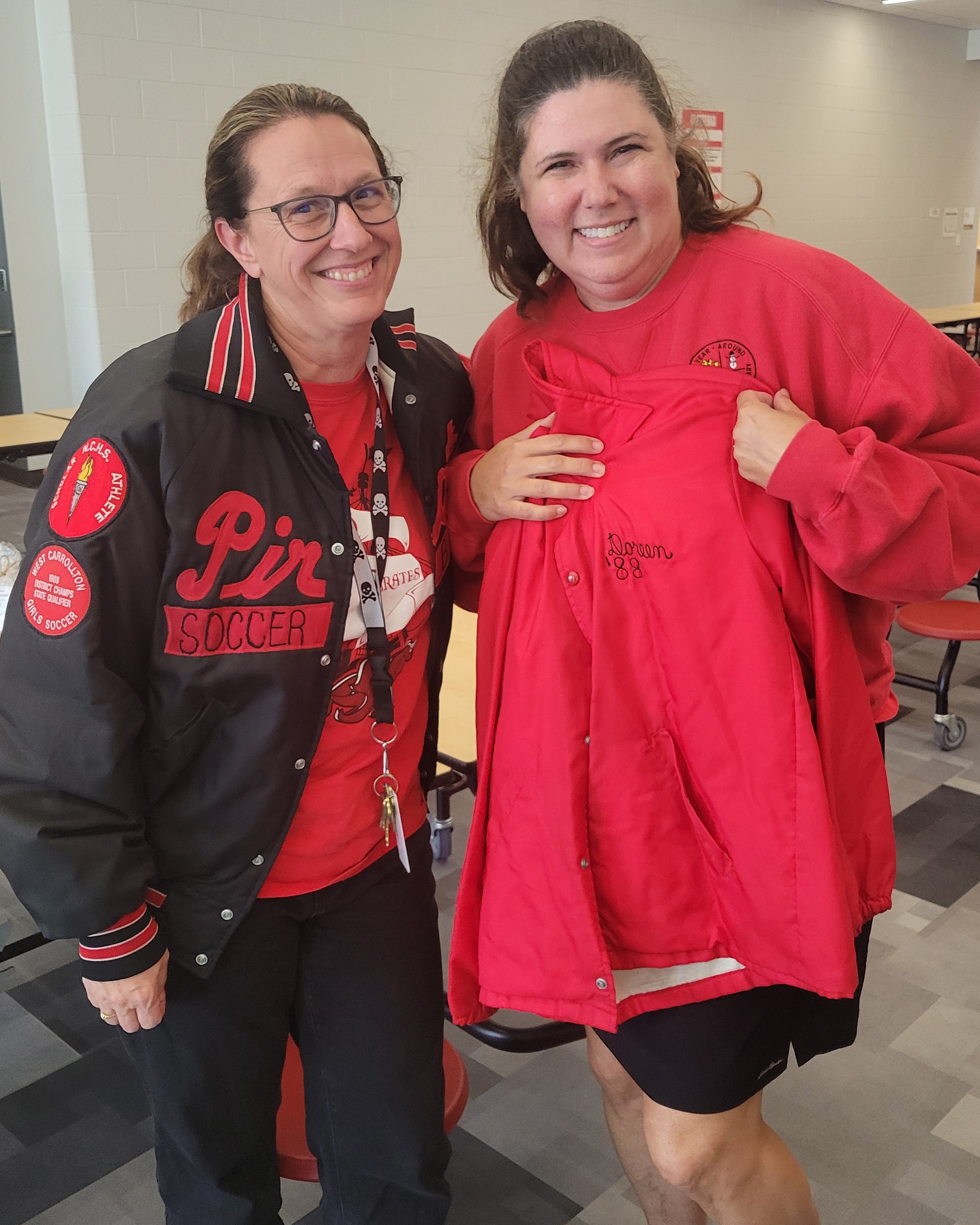 Two women standing in a dining area. One wears a red shirt, and the other wears a red varsity jacket.