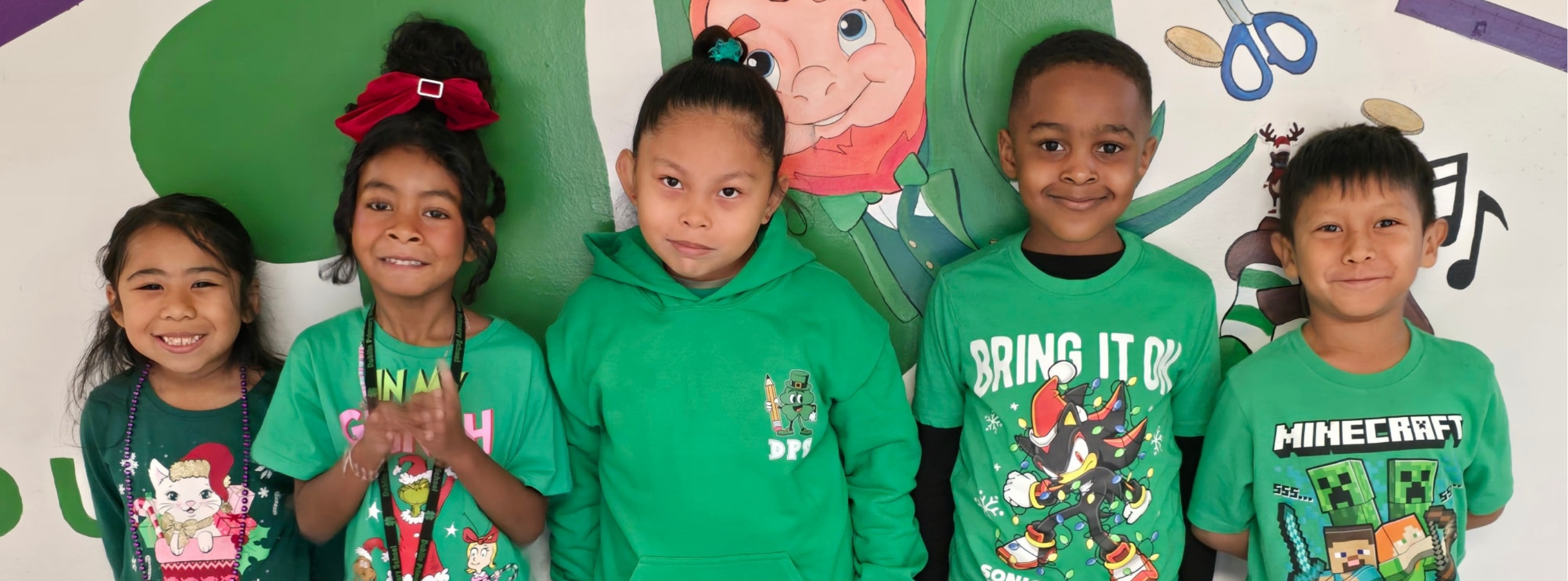 Five elementary students wearing green shirts stand shoulder to shoulder, smiling in front of a colorful school mural featuring playful characters and musical notes.