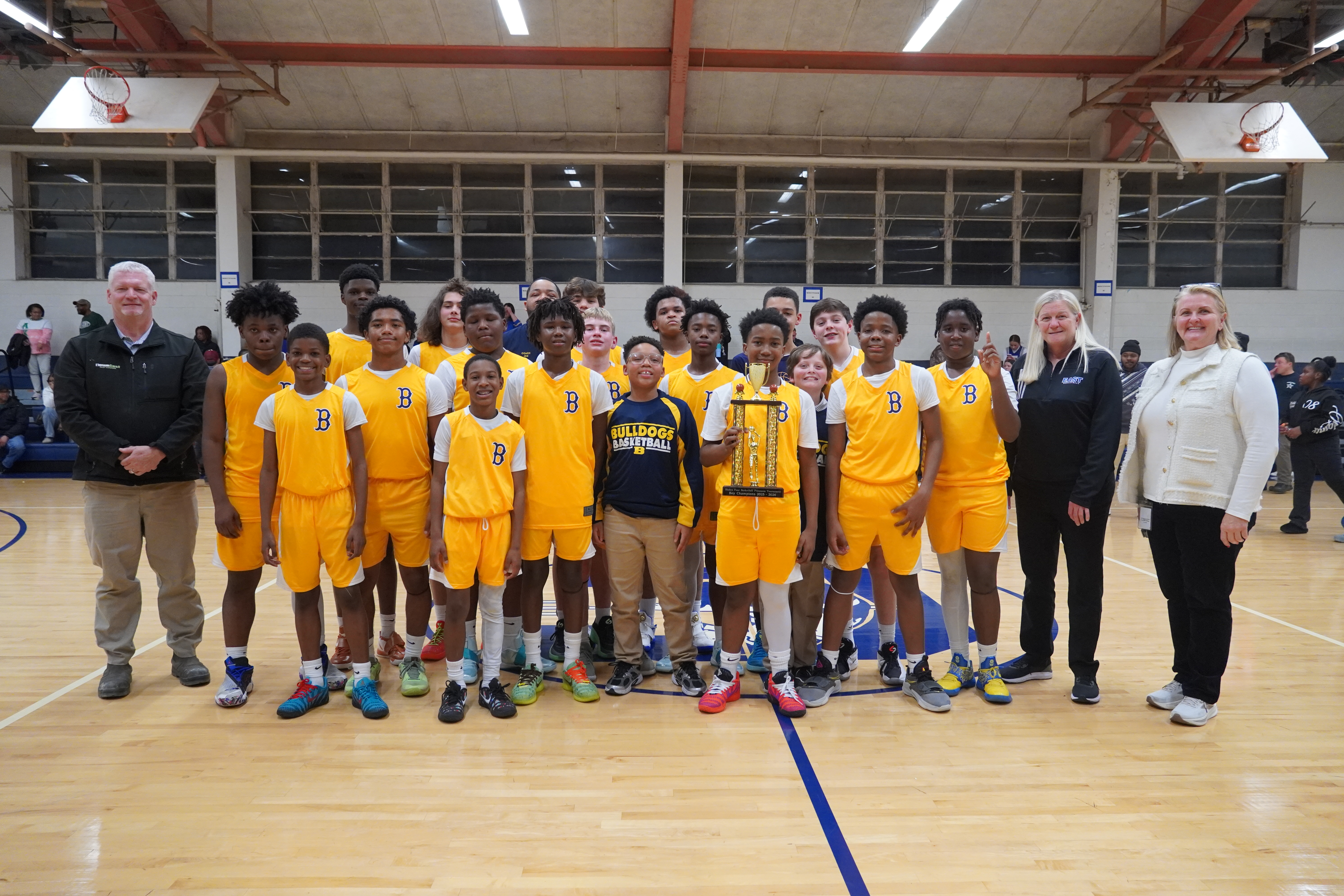 Middle school boys basketball team in gold uniforms poses in a gym with coaches and staff, proudly holding a large championship trophy at center court.