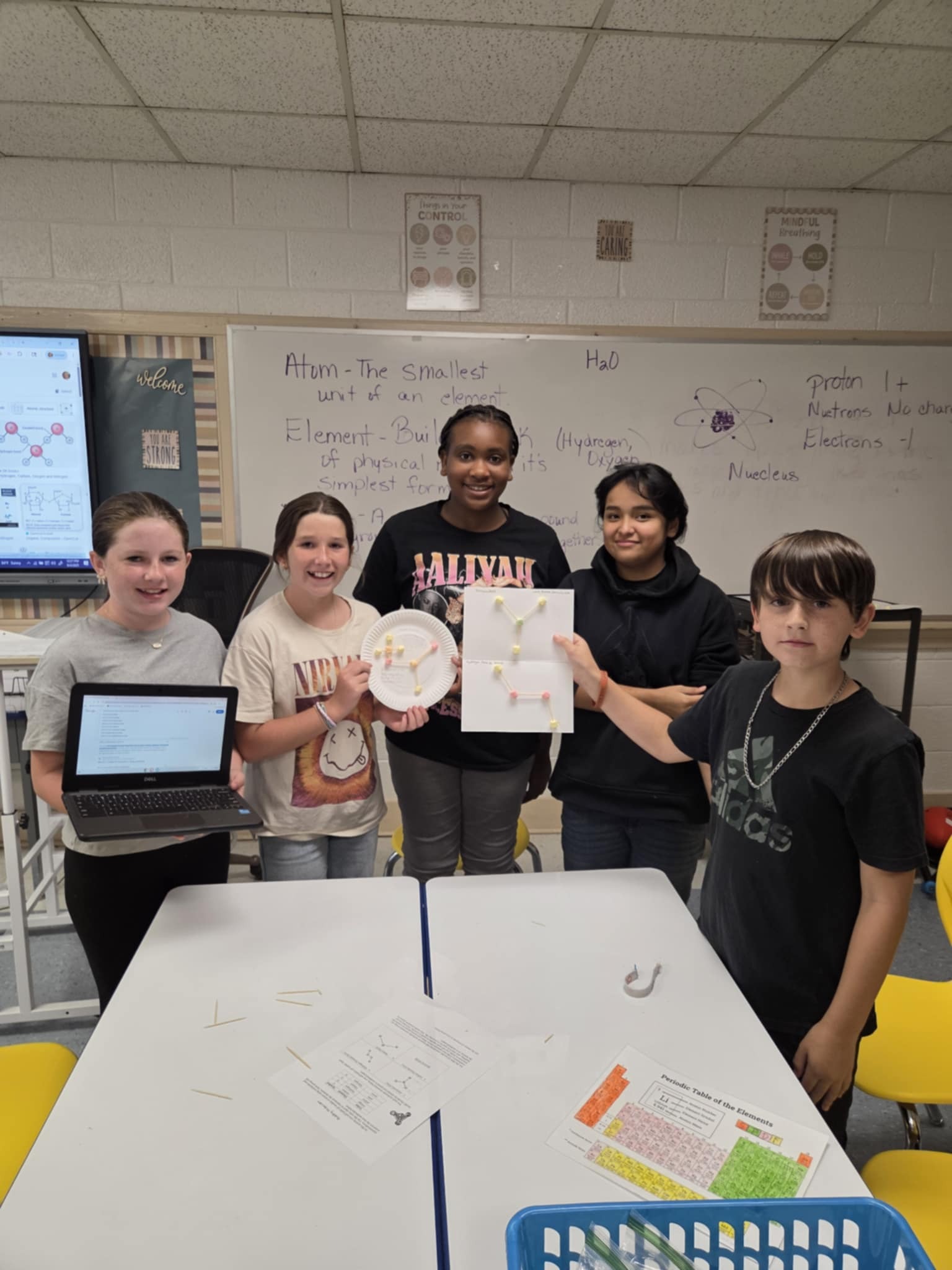 Five students stand in a classroom holding molecule models and a laptop, with a science lesson displayed on the board behind them.