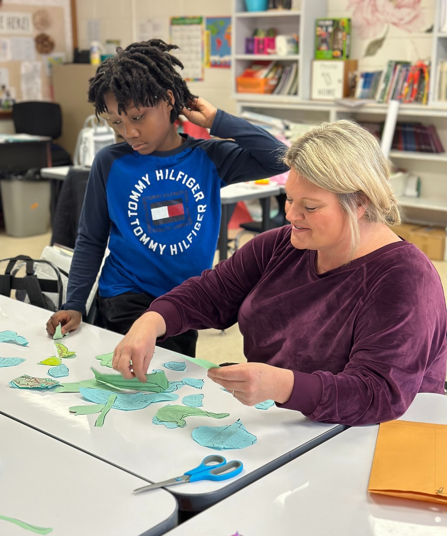 Student and teacher work together at a classroom table, assembling paper leaf cutouts for a collaborative activity.